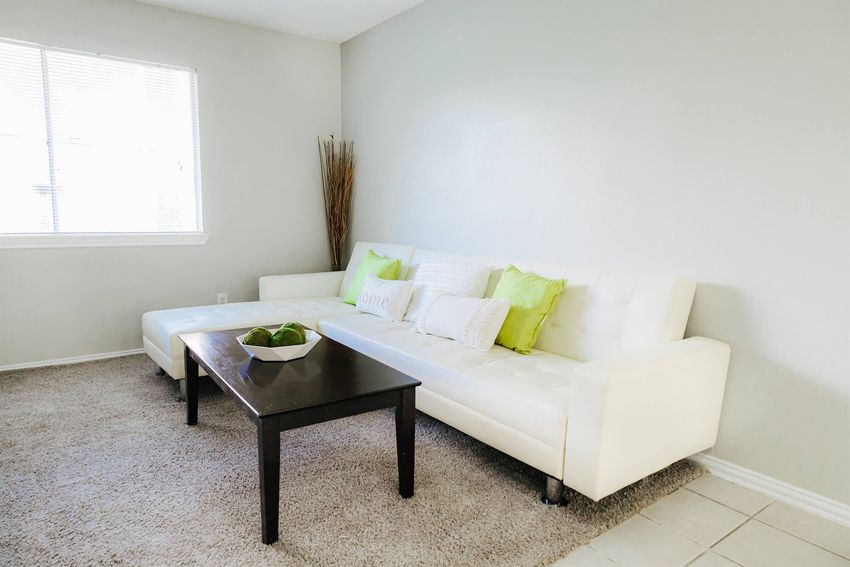 A bright and airy living room featuring a modern white sectional sofa with green accent pillows, a dark wooden coffee table, and a decorative bowl. Natural light flows in through a window, and the floor is covered with light-colored carpet, creating a cozy atmosphere.