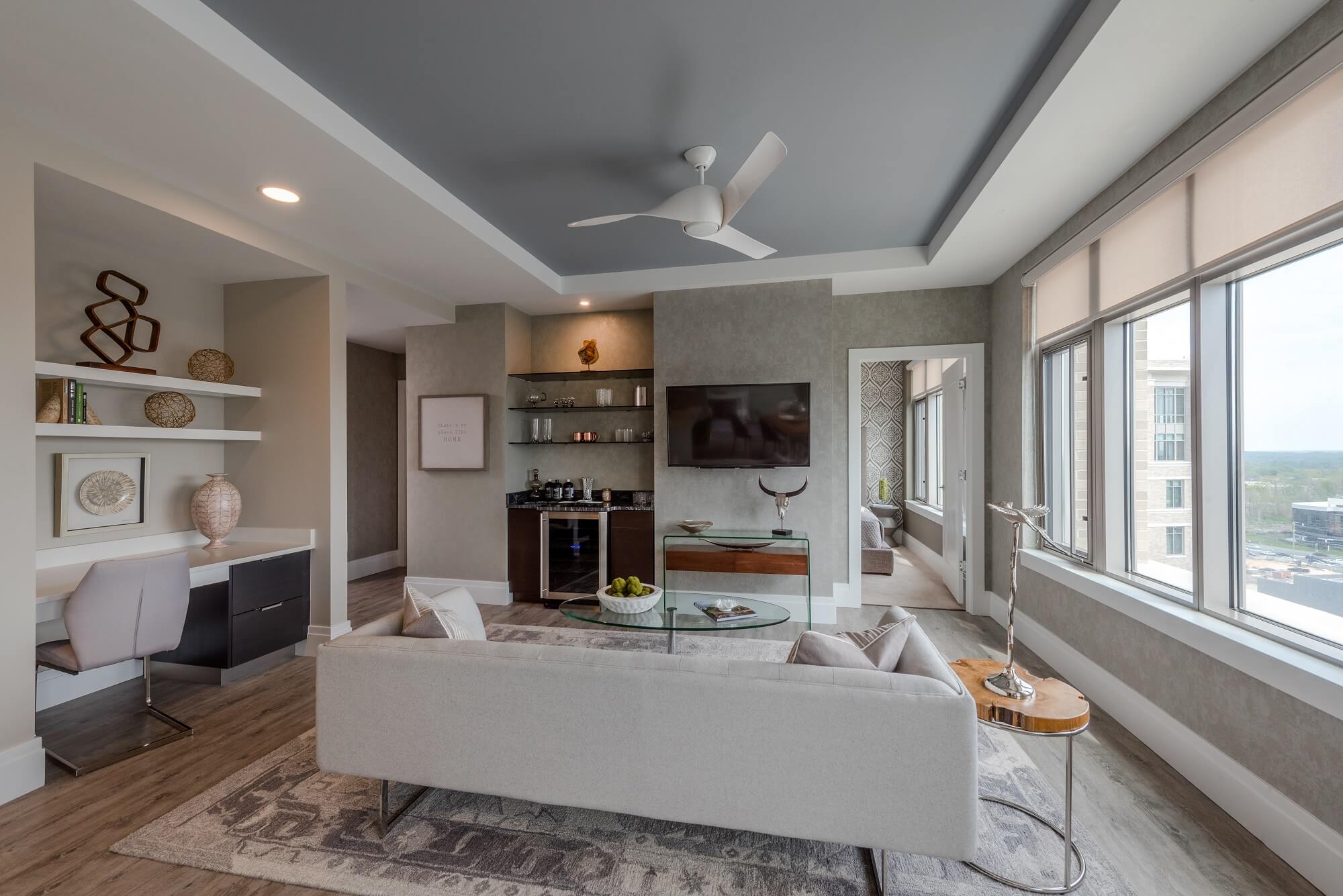 Modern living room with a light gray sofa, glass coffee table, wall-mounted TV, and built-in shelves. Large windows allow natural light in, and a small desk area is tucked into a niche on the left.