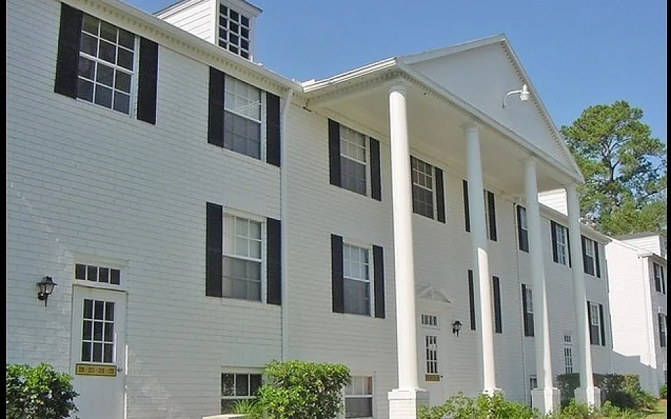 Elegant colonial-style apartment building with grand white pillars and charming black shutters, surrounded by lush greenery and set against a clear blue sky.