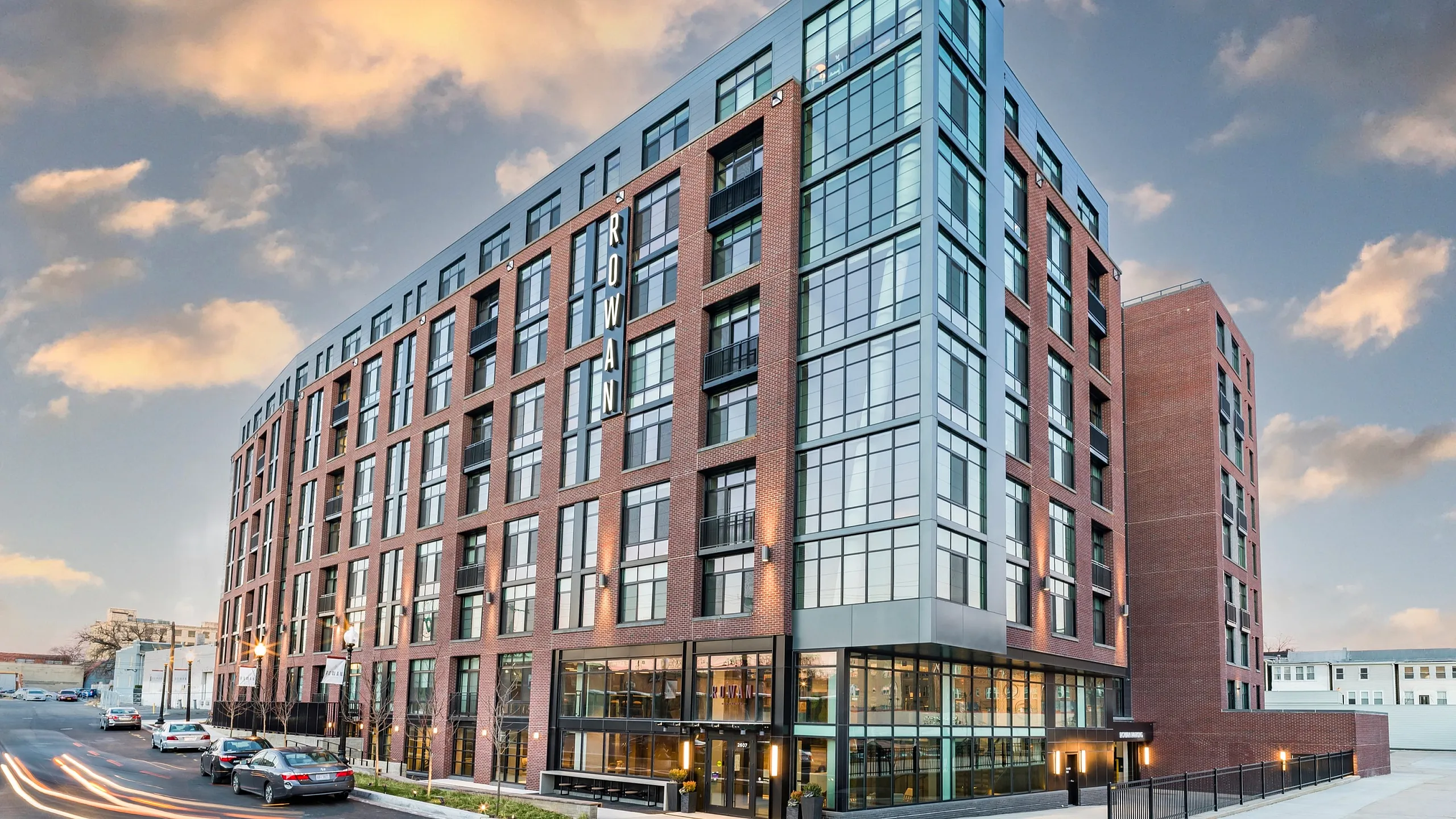 Modern urban apartment building with sleek glass facade and warm brick accents, highlighted against a vibrant sunset sky.