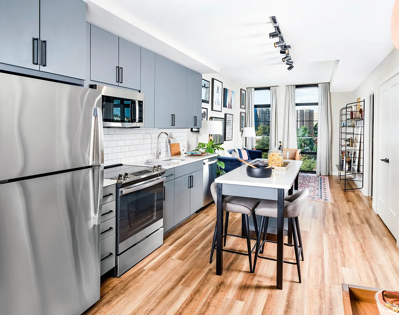 Modern apartment kitchen with sleek gray cabinets, stainless steel appliances, and a cozy dining area leading to a sunlit living room with stylish decor.