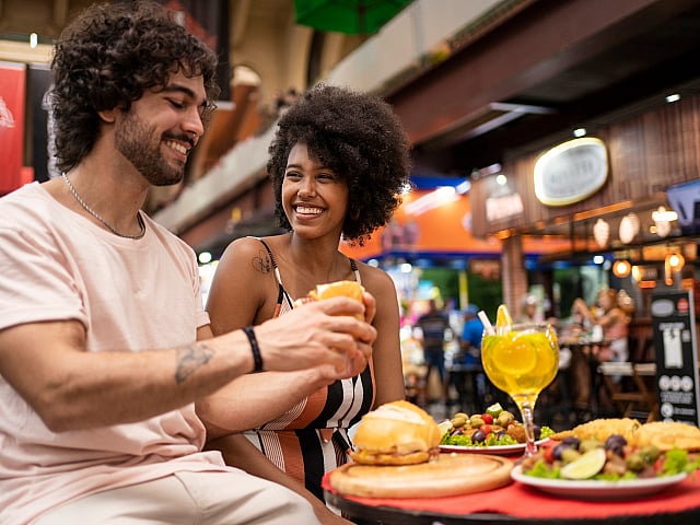 Vibrant market scene with a smiling couple enjoying delicious food and refreshing drinks amidst a lively, colorful backdrop.