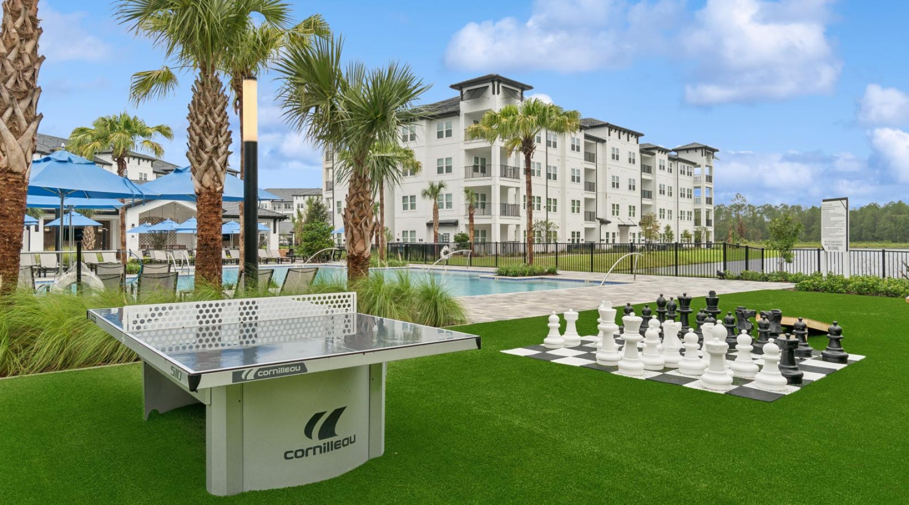 Dog-Friendly Luxury Apartments in St. Johns, FL - Ascend Durbin Park - Poolside Pavilion with Ping-Pong Table and Oversized Chessboard, Trees on a Grassy Lawn, and View of the Pool and Apartments.