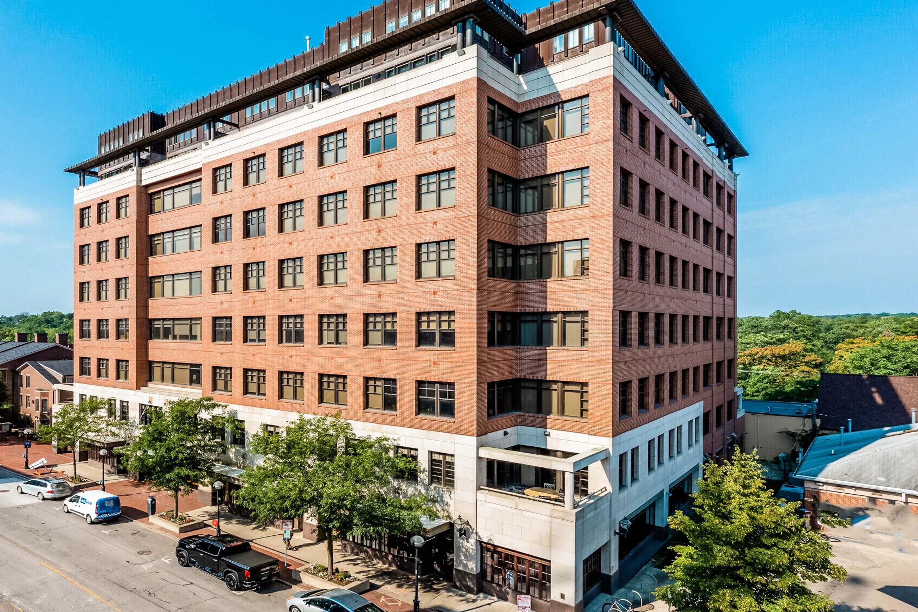 Modern multi-story apartment complex with a red brick facade on a sunny day, street view with parked cars.