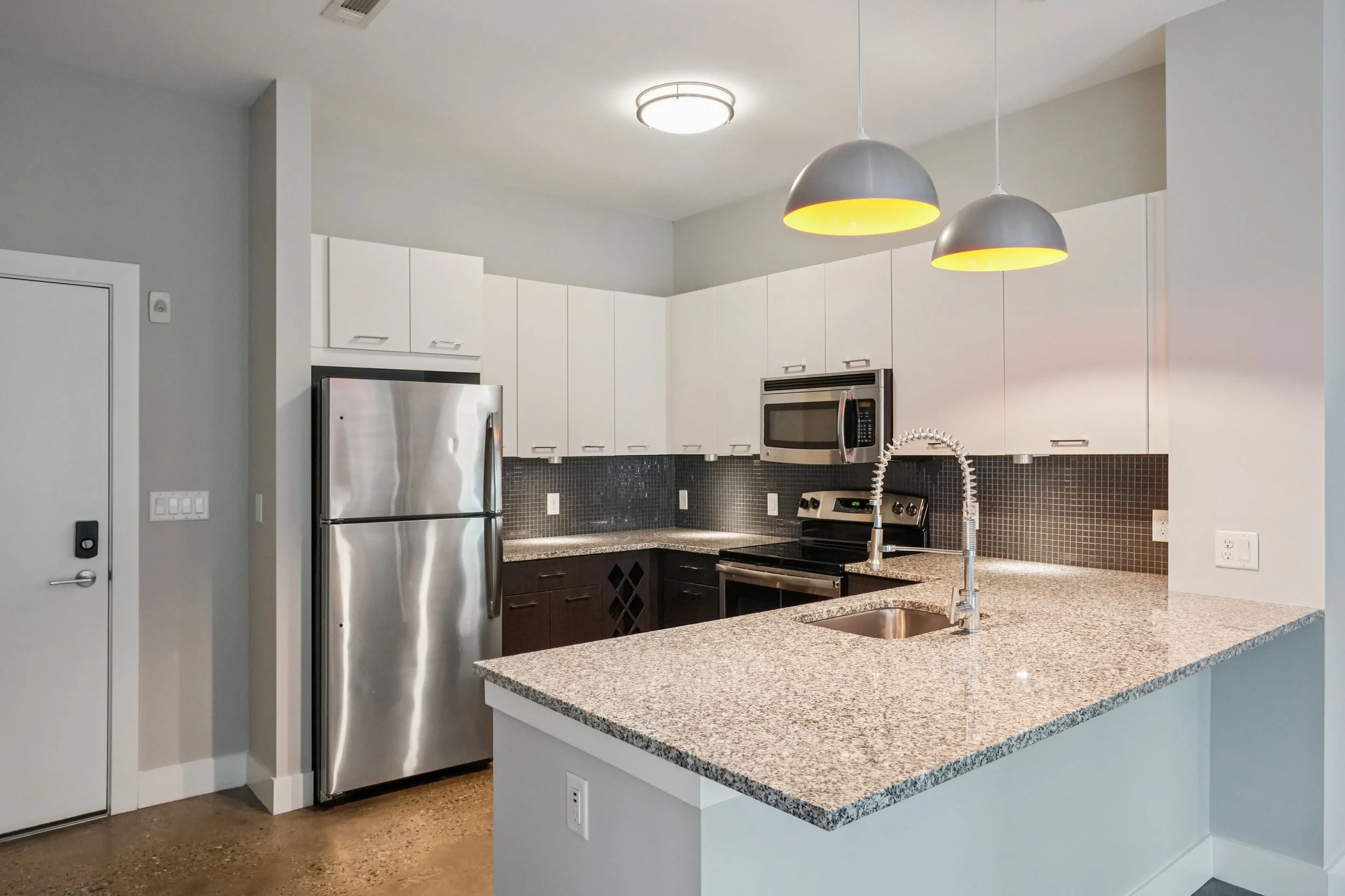 Modern kitchen with white upper cabinets, black lower cabinets, stainless steel appliances including a refrigerator, microwave, and stove, granite countertops, a black tile backsplash, and two gray pendant lights hanging over a countertop with a built-in sink.