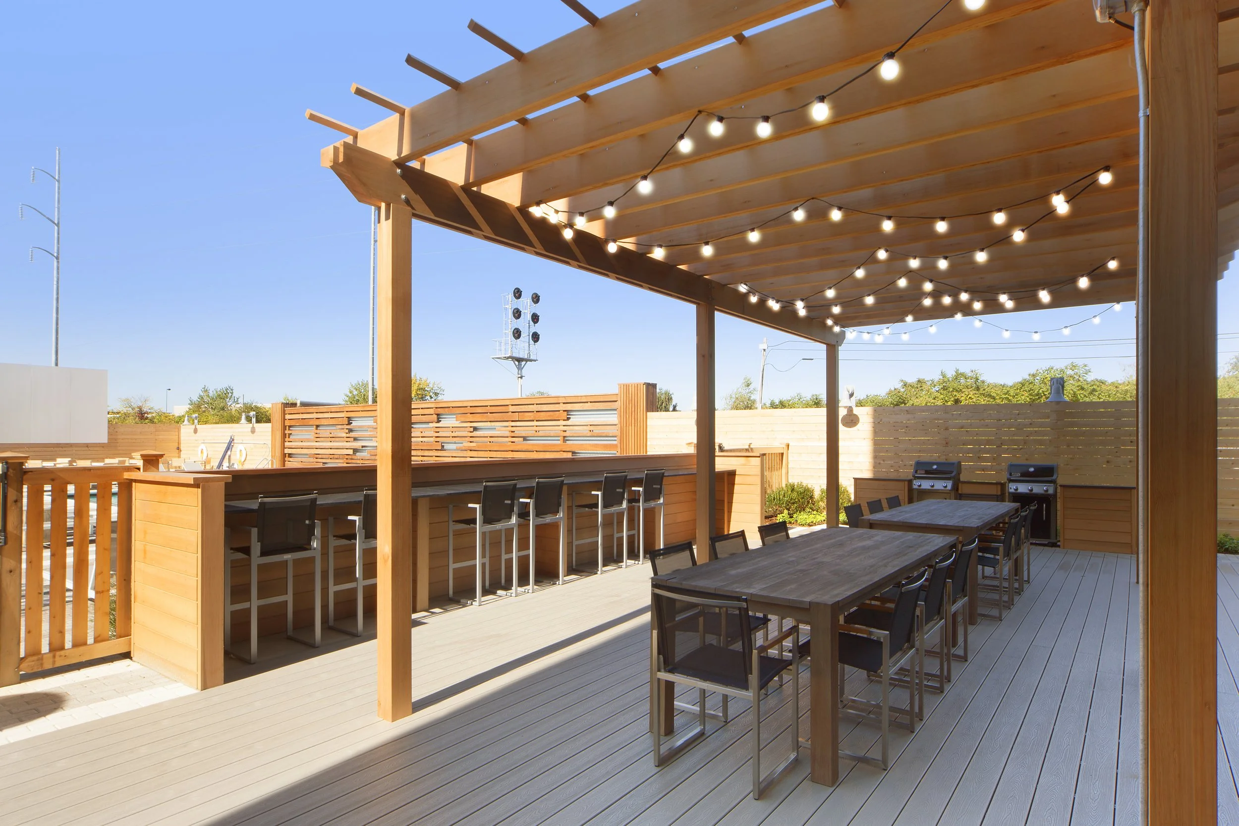 Outdoor patio with wooden pergola, string lights, dining table with chairs, bar area with high stools, and two gas grills, under a partly cloudy sky.