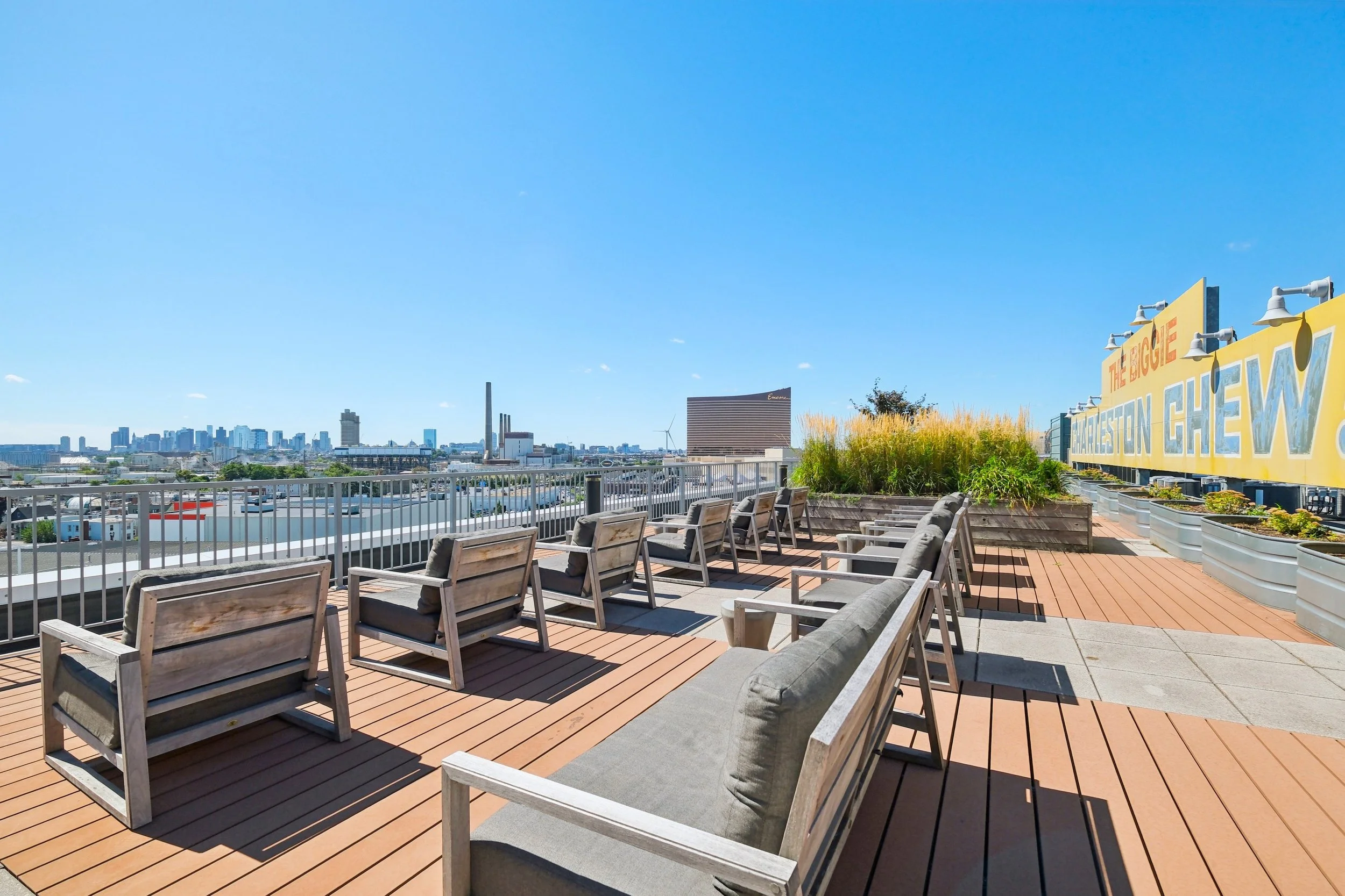 Rooftop patio with wooden decking, seating with cushions, planters with grass and plants, city skyline in the background, clear blue sky.