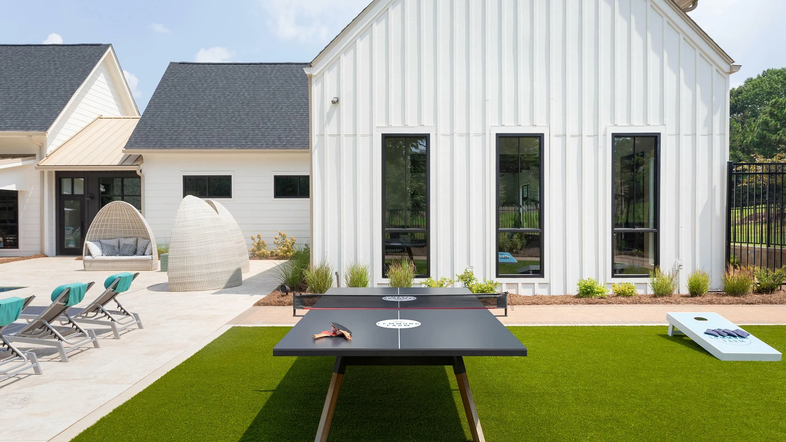 Charming outdoor area featuring a stylish white building with large windows, inviting patio lounges, and a sleek ping-pong table set on vibrant green turf, perfect for leisure and relaxation.