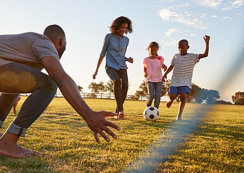 Family enjoying a fun soccer game outdoors at sunset in a spacious green field, promoting a vibrant and active lifestyle.