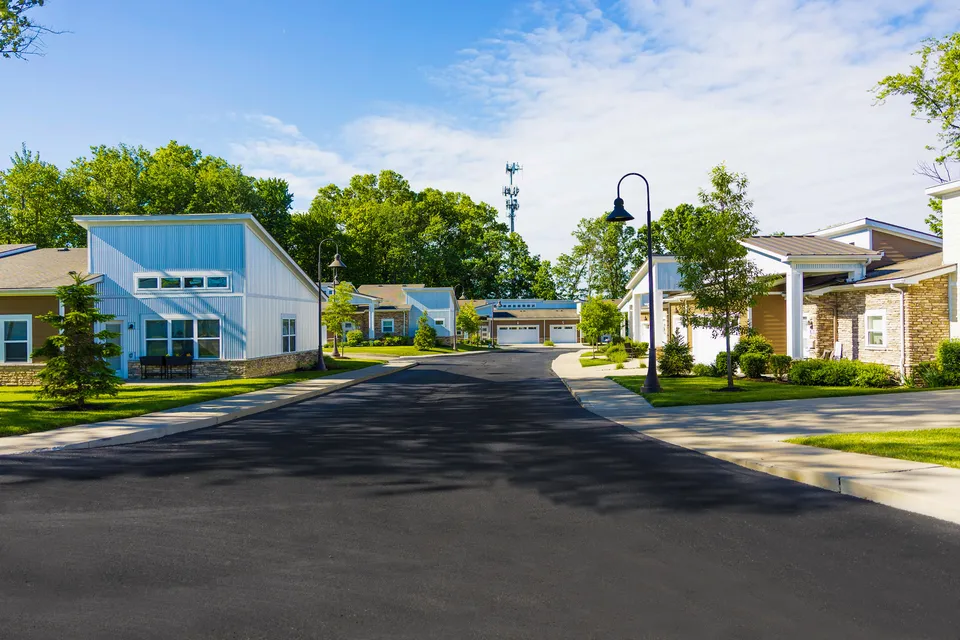 Charming suburban neighborhood with modern single-story homes, manicured lawns, and tree-lined streets under a bright blue sky.