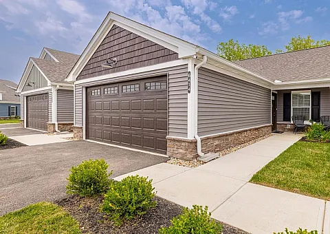 Charming suburban duplex featuring double garages, a manicured front lawn, and inviting front porch seating under a clear blue sky.