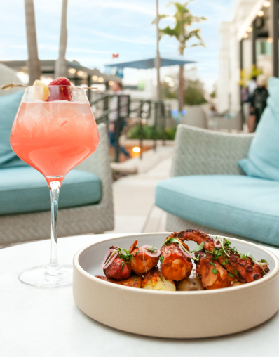 A pink cocktail and a plate of food on a white table in an outdoor restaurant setting.