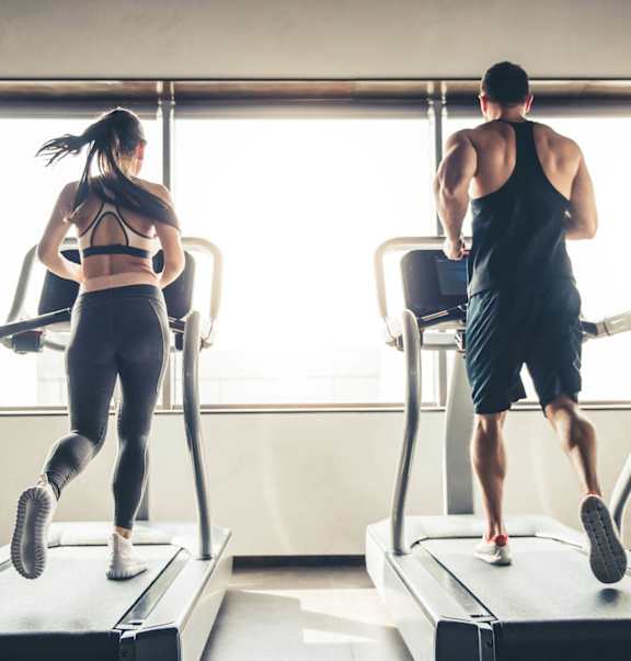 Man and woman running on treadmills in a gym with large windows.
