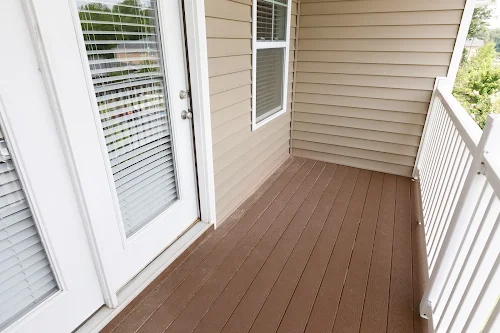 A clean apartment balcony with brown decking, sliding glass doors, and light-colored siding.
