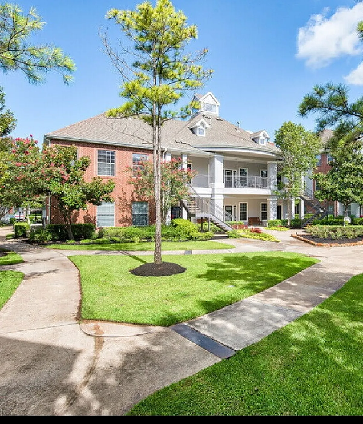 Charming apartment complex with lush greenery, elegant brick buildings, and serene walkways under a bright blue sky.