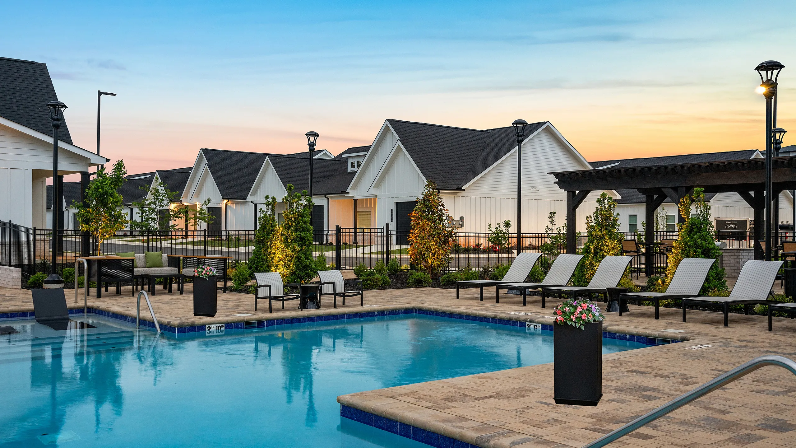 Charming community area featuring a sparkling swimming pool surrounded by elegant lounge chairs and lush landscaping at sunset.