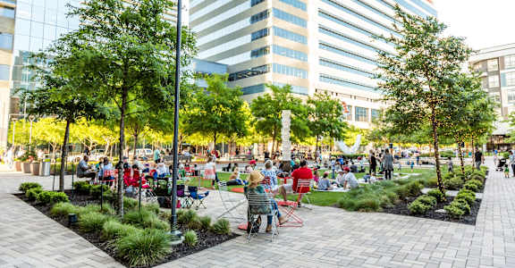 A park with people sitting and standing on the grass and pavement.