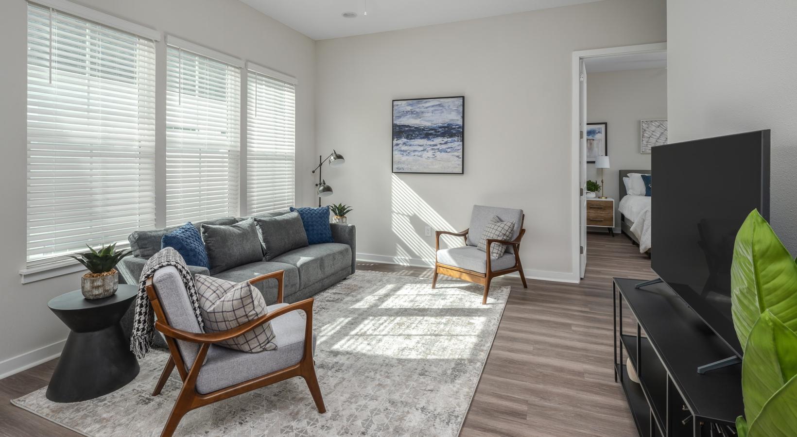 Jacksonville, FL, Townhomes Near Ponte Vedra Beach - Cottage Street at eTown - Bright Living Room With Large Windows, Gray Sofa, Accent Chairs, Side Table, Ceiling Fan, and View Into Bedroom.
