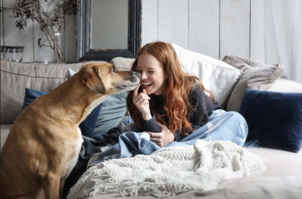 A woman sits on a couch, gently petting a dog resting beside her, both appearing relaxed and content.