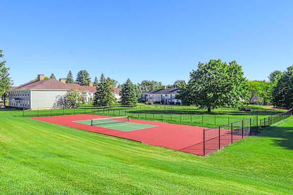 A tennis court is surrounded by a green lawn and a fence.
