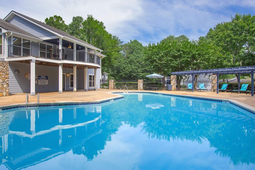 A clear blue swimming pool surrounded by a stone patio, with a two-story building in the background featuring a covered balcony. Lush green trees and a pergola provide shade, and several blue lounge chairs are positioned around the pool area under a blue sky with scattered clouds.
