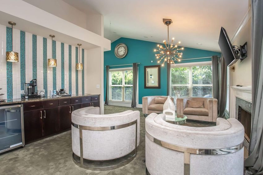 A modern living space featuring a cozy seating area with two circular armchairs and a beige sofa, complemented by a stylish chandelier and wall clock. The decor includes textured blue and white wallpaper, large windows with natural light, and a sleek wet bar with a mini fridge.