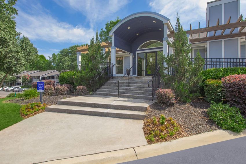 A welcoming entrance to a modern building with a curved roof, surrounded by greenery and well-maintained landscaping. There are stone steps leading up to the door, with handrails on either side. Clear blue skies overhead add to the bright atmosphere of the scene.