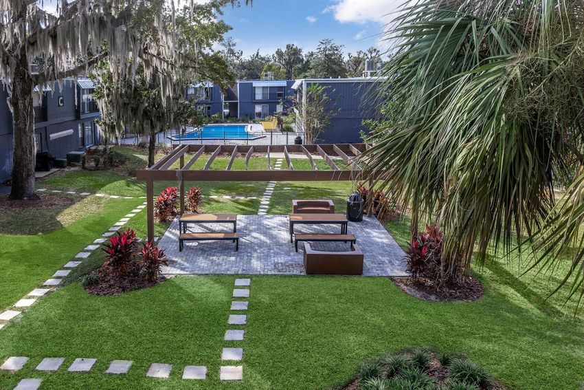 A landscaped outdoor area featuring a pergola over seating arrangement with modern benches. It is surrounded by lush green grass, decorative plants, and stepping stone pathways. In the background, a swimming pool is visible amidst apartment buildings, creating a tranquil recreational space.