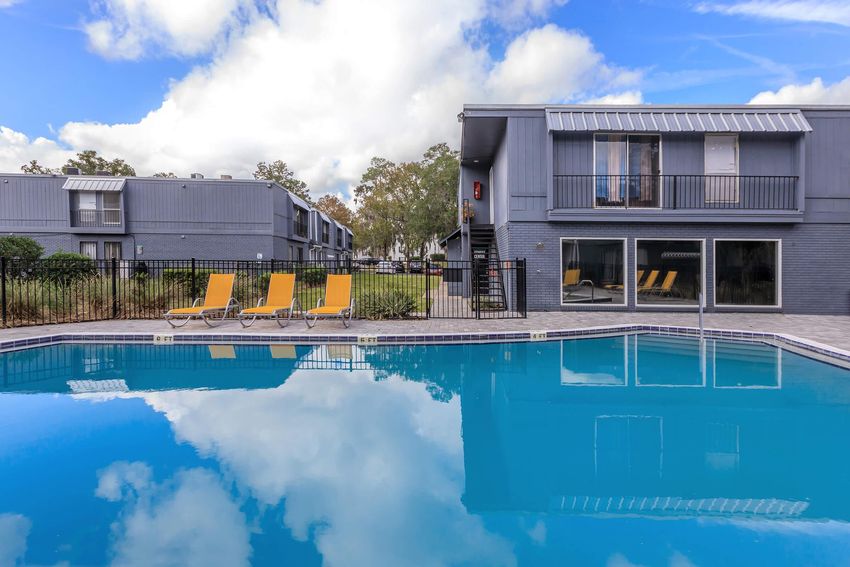 A clear blue swimming pool reflects the sky and surrounding buildings. Yellow lounge chairs are positioned near the water, and gray apartment buildings with balconies are visible in the background. The scene is set against a partly cloudy sky, creating a tranquil and inviting atmosphere.