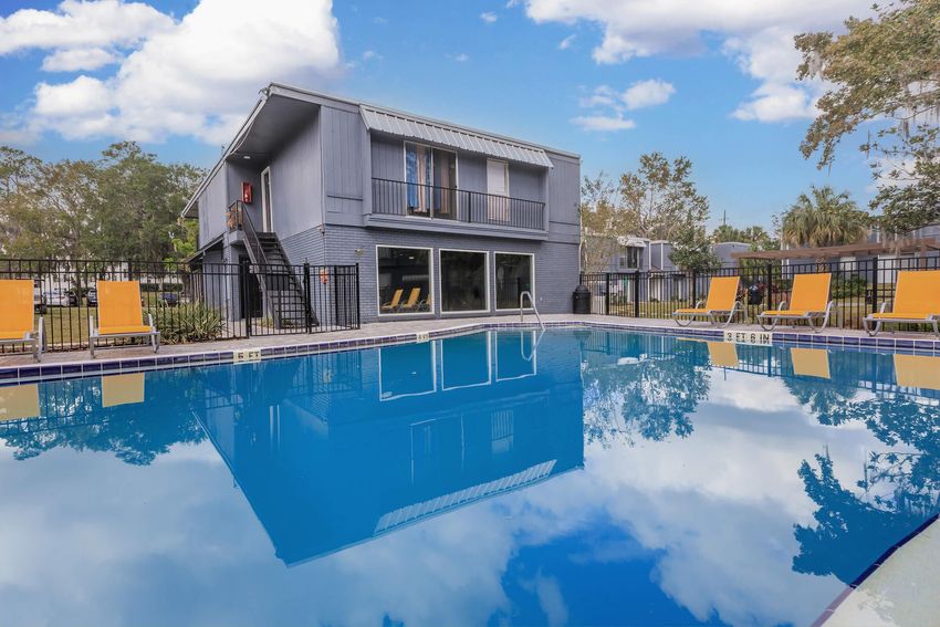 A clear blue swimming pool reflecting clouds and a nearby building. The pool area features bright yellow lounge chairs arranged around it, with greenery and trees in the background, creating a serene outdoor environment. The setting appears inviting and well-maintained.