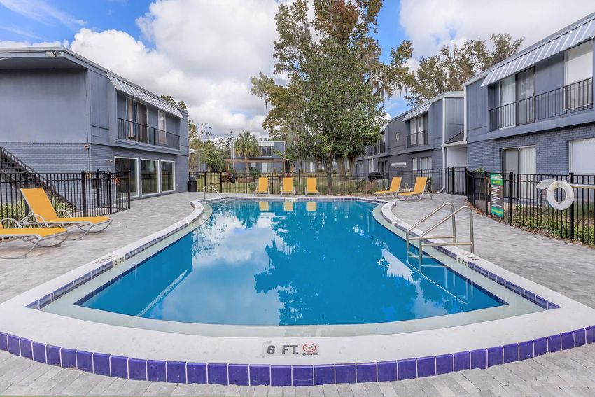 A swimming pool surrounded by yellow lounge chairs and a spacious patio area. The pool is marked with a "6 FT" depth sign, indicating the water level. Two buildings with gray exteriors are visible in the background, along with trees and a partly cloudy sky. The area is fenced for safety.