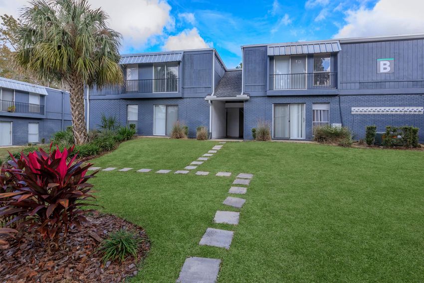 A well-maintained lawn featuring a stone pathway leading to a two-story apartment building. The building has gray exterior walls with balconies, surrounded by vibrant green grass and tropical plants. The sky is partly cloudy, creating a bright and inviting atmosphere.