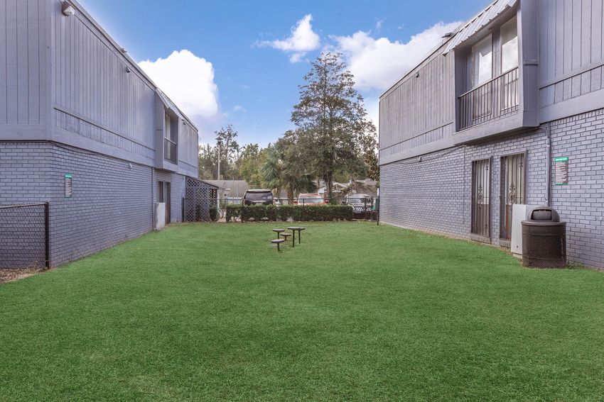A view of a small open grassy area between two gray residential buildings. There are a few picnic tables on the grass, surrounded by low shrubs and a fence. The sky is blue with some clouds, creating a tranquil outdoor environment.