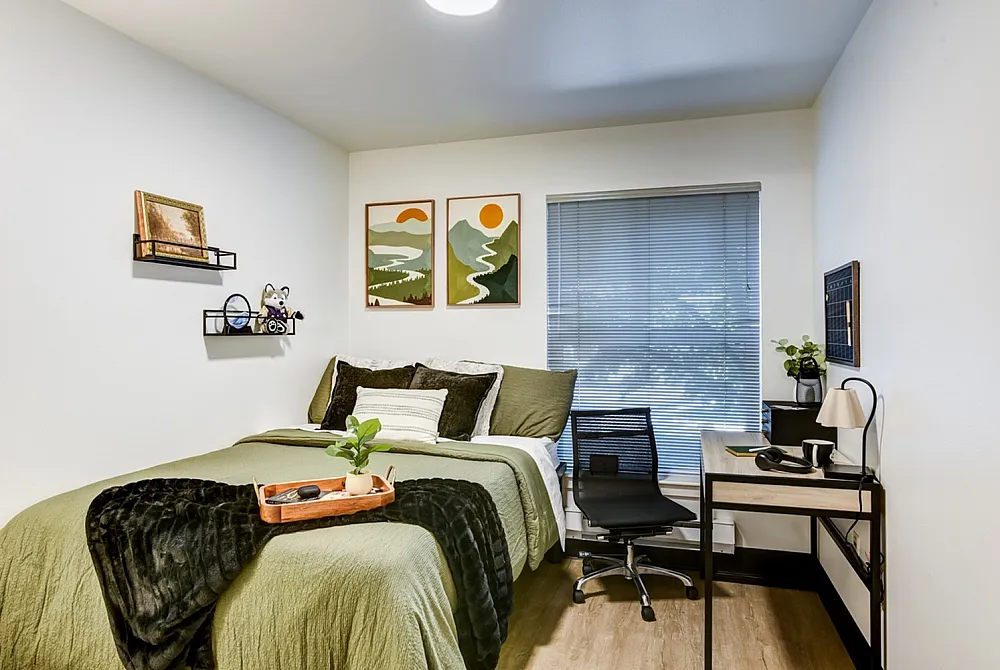 Inviting bedroom with modern decor, featuring a cozy bed with green tones, a sleek desk setup with chair, and stylish wall art.