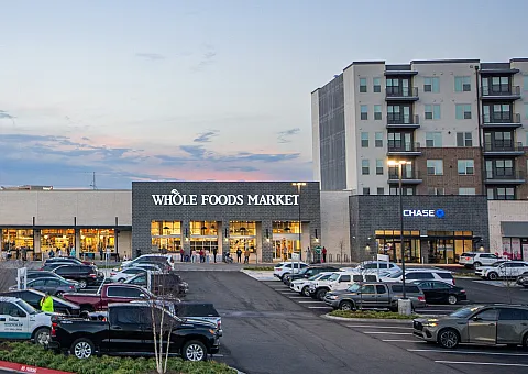 Contemporary retail complex at dusk featuring Whole Foods Market, ample parking, and modern apartments, offering convenience and style.