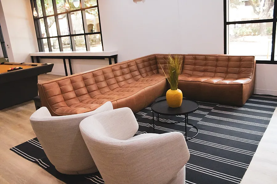 Cozy living area with a stylish brown sectional sofa, elegant beige chairs, and a vibrant yellow vase on a modern coffee table, all set on a chic black and white striped rug near a pool table.