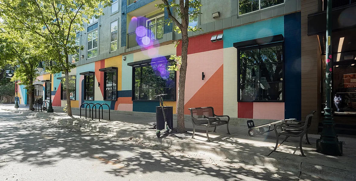 Vibrant city street view with modern apartments featuring colorful geometric murals, lush greenery, and inviting outdoor seating.