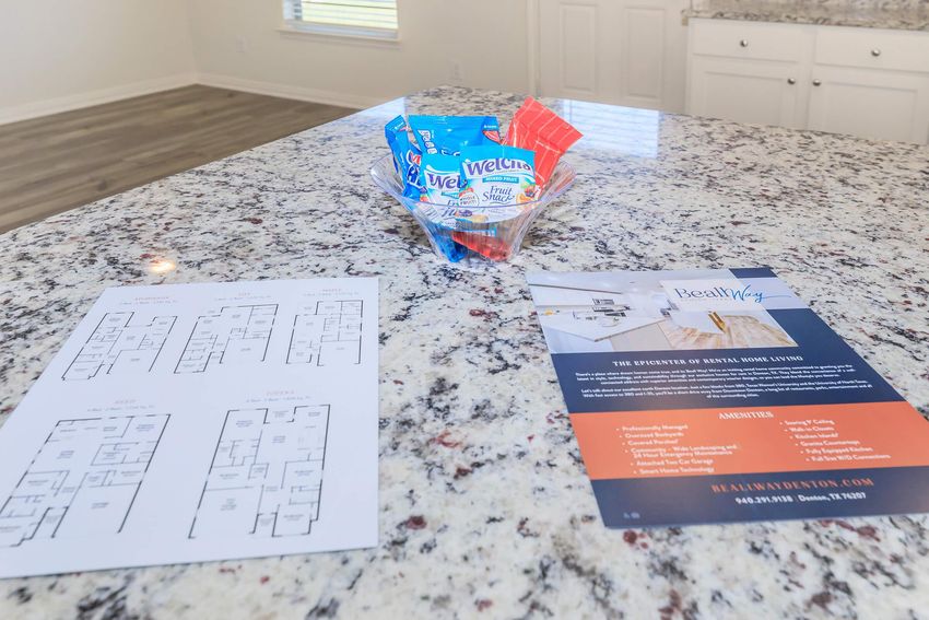 A countertop featuring a brochure for a rental property, a floor plan layout sheet, and a dish of candy. The setting suggests a welcoming environment, possibly in a model home or rental office, with modern decor and natural lighting.
