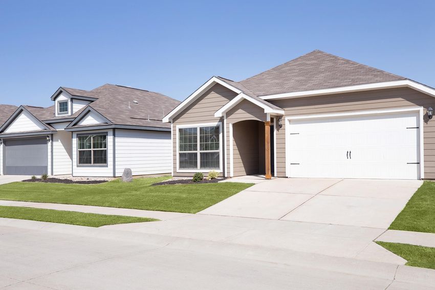 Two residential houses in a suburban neighborhood. The house on the left features a light-colored exterior with a gabled roof, while the house on the right has a darker brown facade and a double garage. Both homes have well-maintained lawns and a clear blue sky in the background.