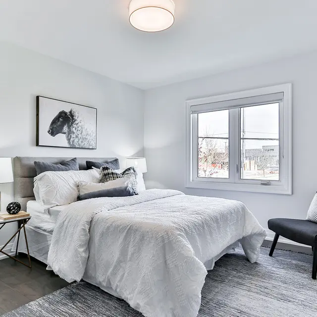 Cozy bedroom with a bed, nightstand, lamp, and a chair near a window, featuring neutral tones and modern decor.