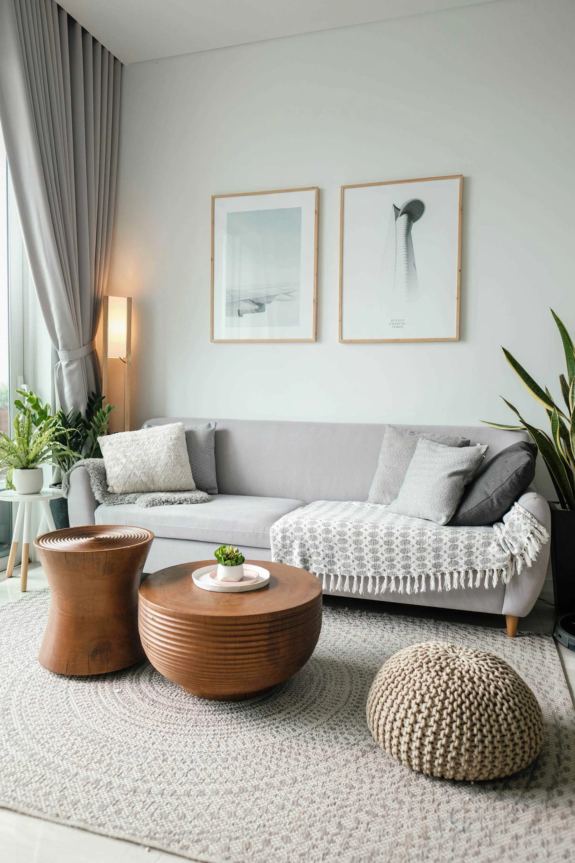 A modern living room featuring a gray sofa with a knit throw, wooden coffee table, and decorative plants. Wall art includes two framed minimalist prints. There is a cozy atmosphere created by soft furnishings and natural light.