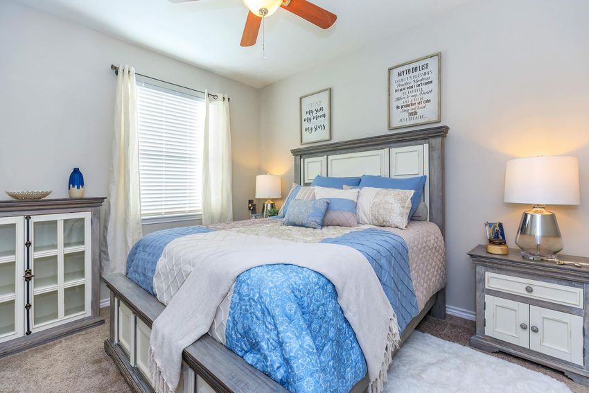 Bright and cozy bedroom featuring a large bed with blue and white bedding, decorative pillows, and a matching throw. There's a nightstand with a lamp on either side of the bed, a glass-front cabinet in the corner, and framed wall art above the bed, all contributing to a warm and inviting atmosphere. Natural light filters in through the windows.