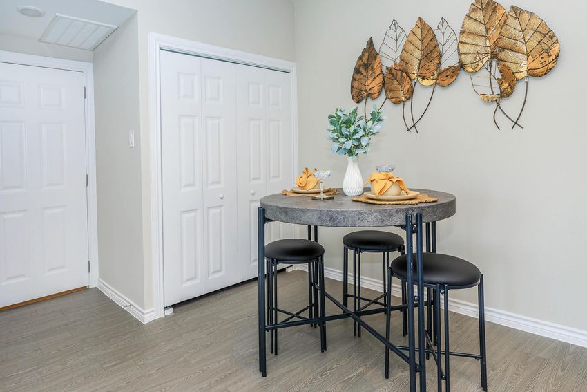 A cozy dining area featuring a round concrete table with three black stools. The table is set with a beige table runner and yellow napkins. A decorative arrangement of leaves is mounted on the wall above, and a small vase with greenery is placed on the table. Two white doors are visible in the background.