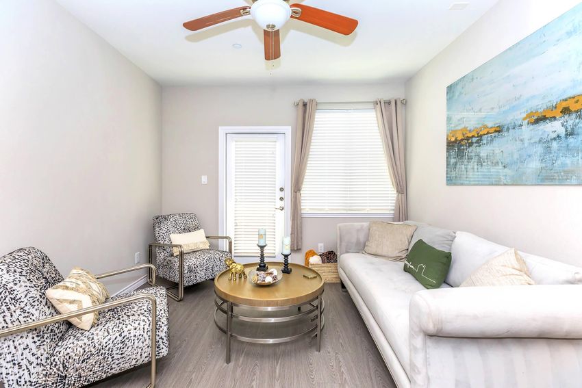 A modern living room featuring a light gray color scheme. There is a cozy sofa and two patterned chairs surrounding a round coffee table. Natural light comes through a nearby window and a glass door leads outside. Decorative elements include a small vase and artwork on the wall.