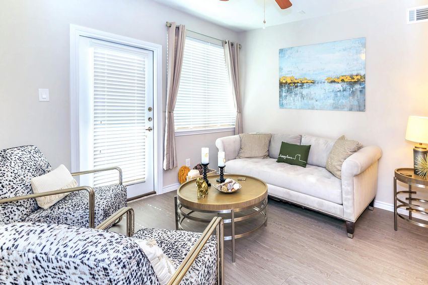 A cozy living room featuring a light-colored sofa with decorative cushions, two patterned armchairs, a round coffee table with candles and decor, a piece of art on the wall, and a window with blinds letting in natural light. The overall color scheme is soft and inviting, creating a warm atmosphere.