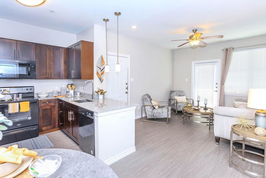 Modern kitchen and living area featuring a countertop with bar stools, dark wood cabinetry, stainless steel appliances, and a cozy seating arrangement. A ceiling fan, natural light from windows, and decorative accents create a welcoming atmosphere.