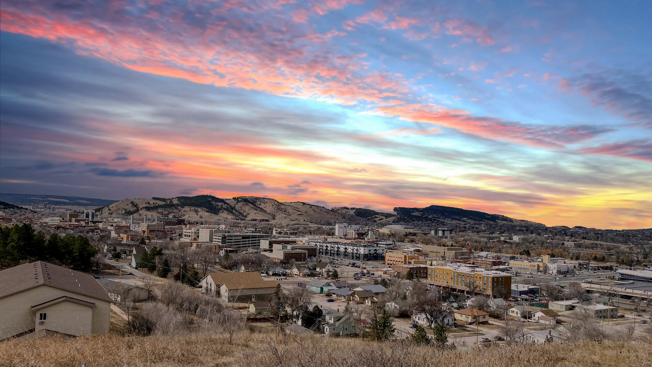 Stunning panoramic view of a vibrant cityscape at sunset, nestled between rolling hills and under a colorful, dramatic sky.