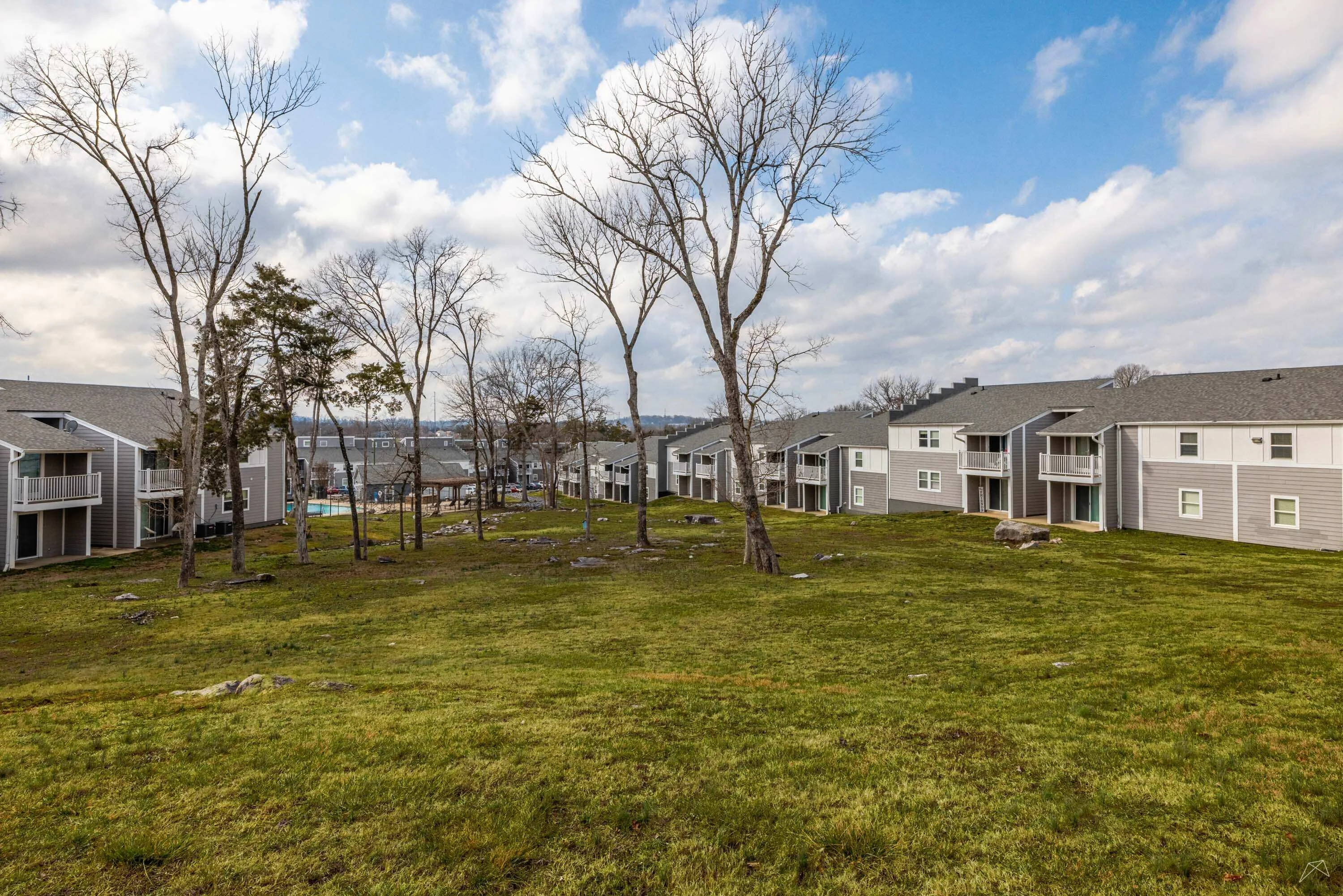 A view of an apartment complex with gray buildings, trees, and a grassy area under a cloudy sky.