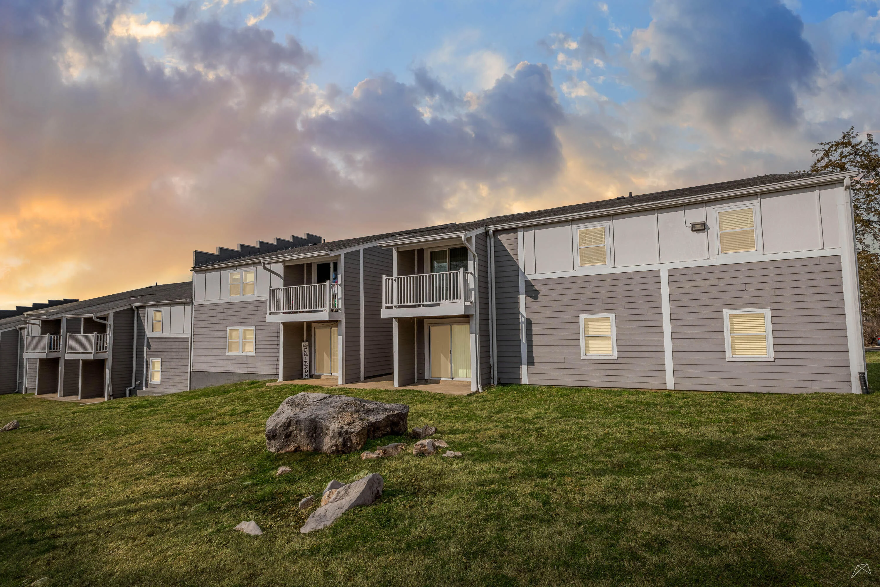 Exterior view of a apartment with multiple units, featuring balconies and a grassy area under a cloudy sky.