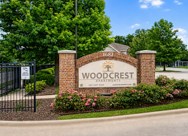The image shows a sign for Woodcrest Apartments with a black fence and flowers in the foreground.